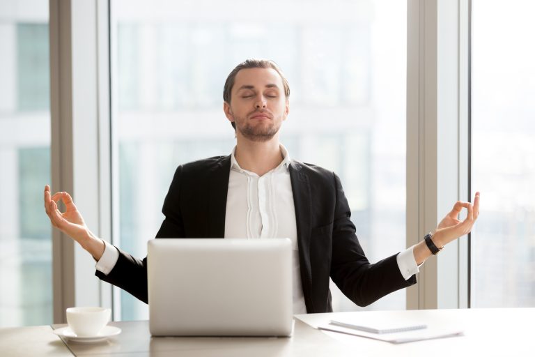 Relaxed young entrepreneur meditating at desk in front of laptop. Office worker doing yoga exercises at workplace. Successful businessman relieves stress with spiritual practices or autosuggestion
